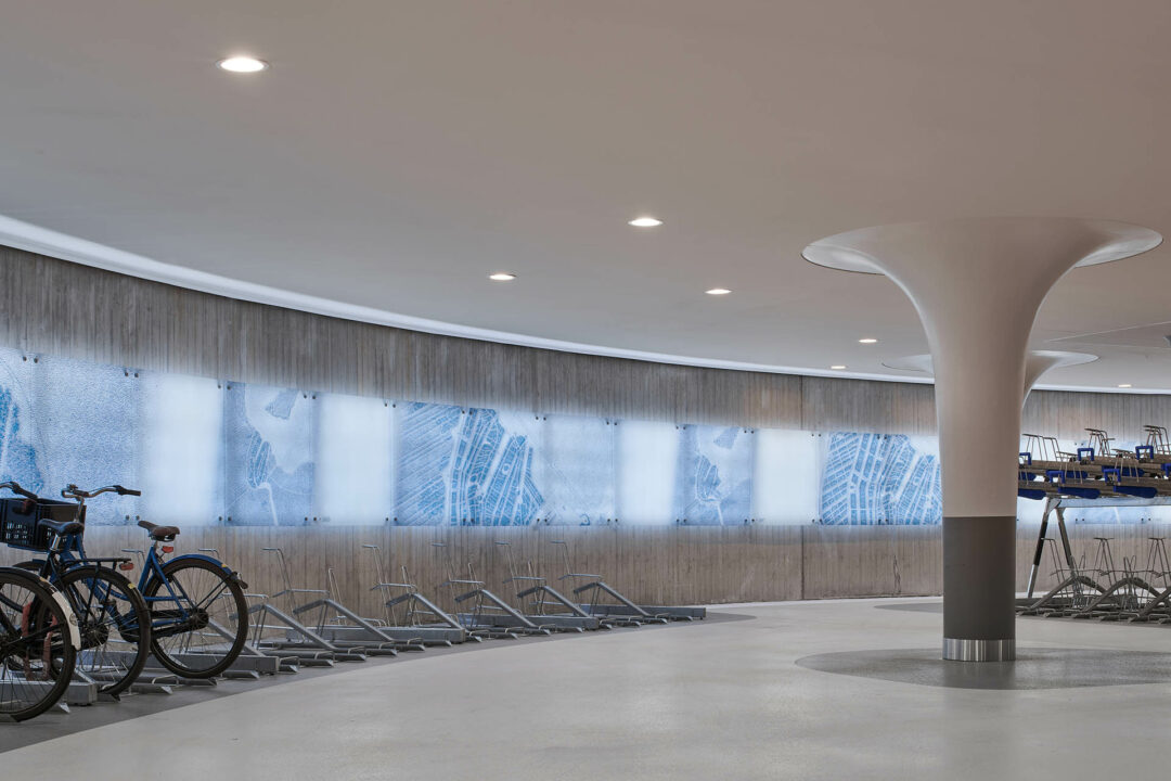 Foto in de nieuwe ondergrondse fietsenstalling in Amsterdam, met de focus op een rij lichtgevende panelen aan de wand. Op de panelen zie je door een algoritme gemaakte kruising tussen groeikaarten van Amsterdam en beelden uit het archief van het Amsterdam Museum.
